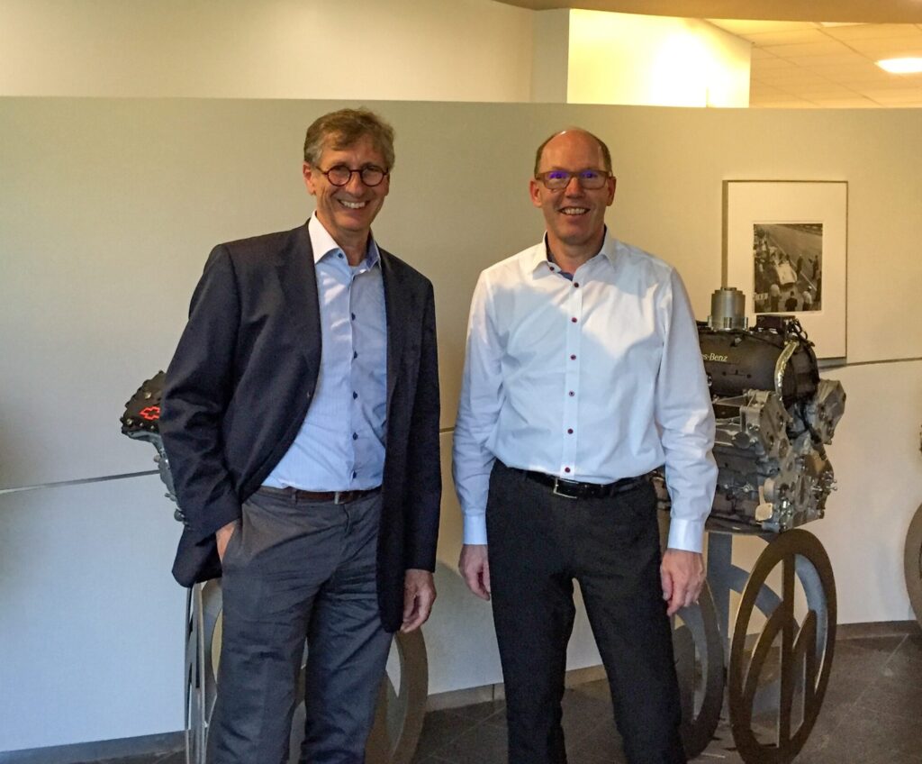 Mario Illien (left) and Stefan Battlogg (right) standing together in front of high-performance engine components, both smiling at the camera.